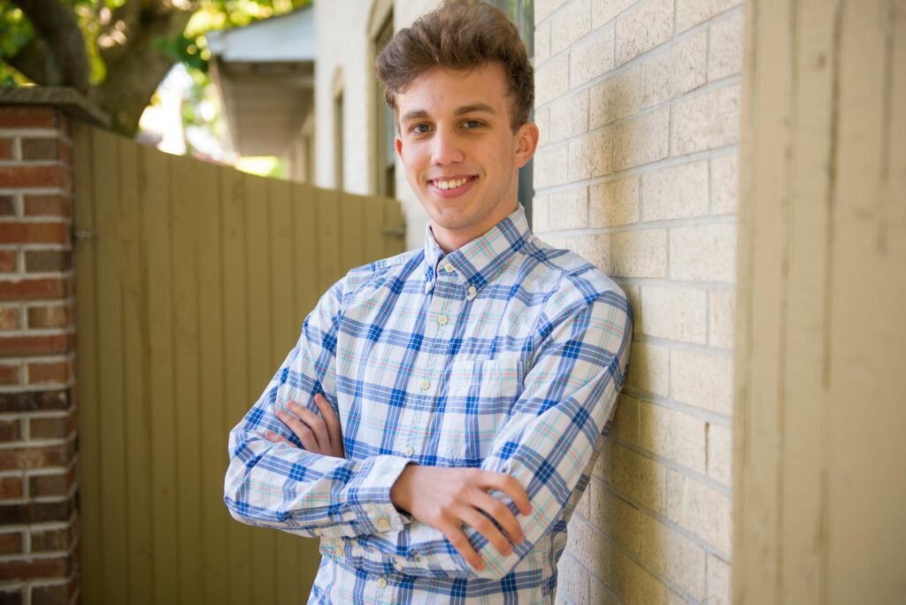 Casually dressed young man smiling, leaning against a brick wall outdoors on a sunny day.